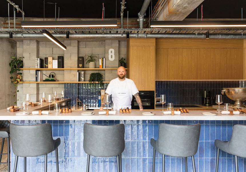 Chef Karl Martin-Boulton stands smiling behind his kitchen counter which is fronted with blue tiles and grey bar stools