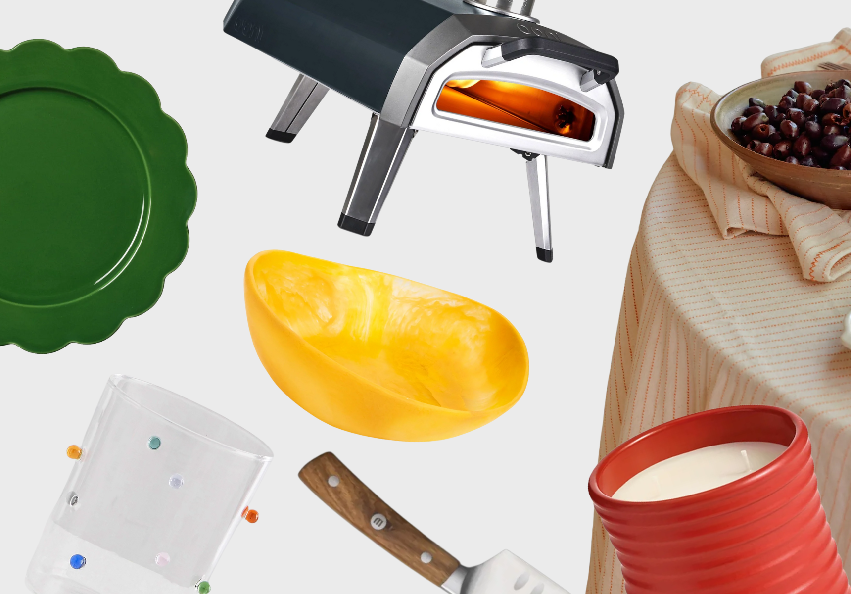 A pizza oven, tablecloth, dinner plates, serving bowl, drinking glasses, a set of knives and a tomato-scented candle